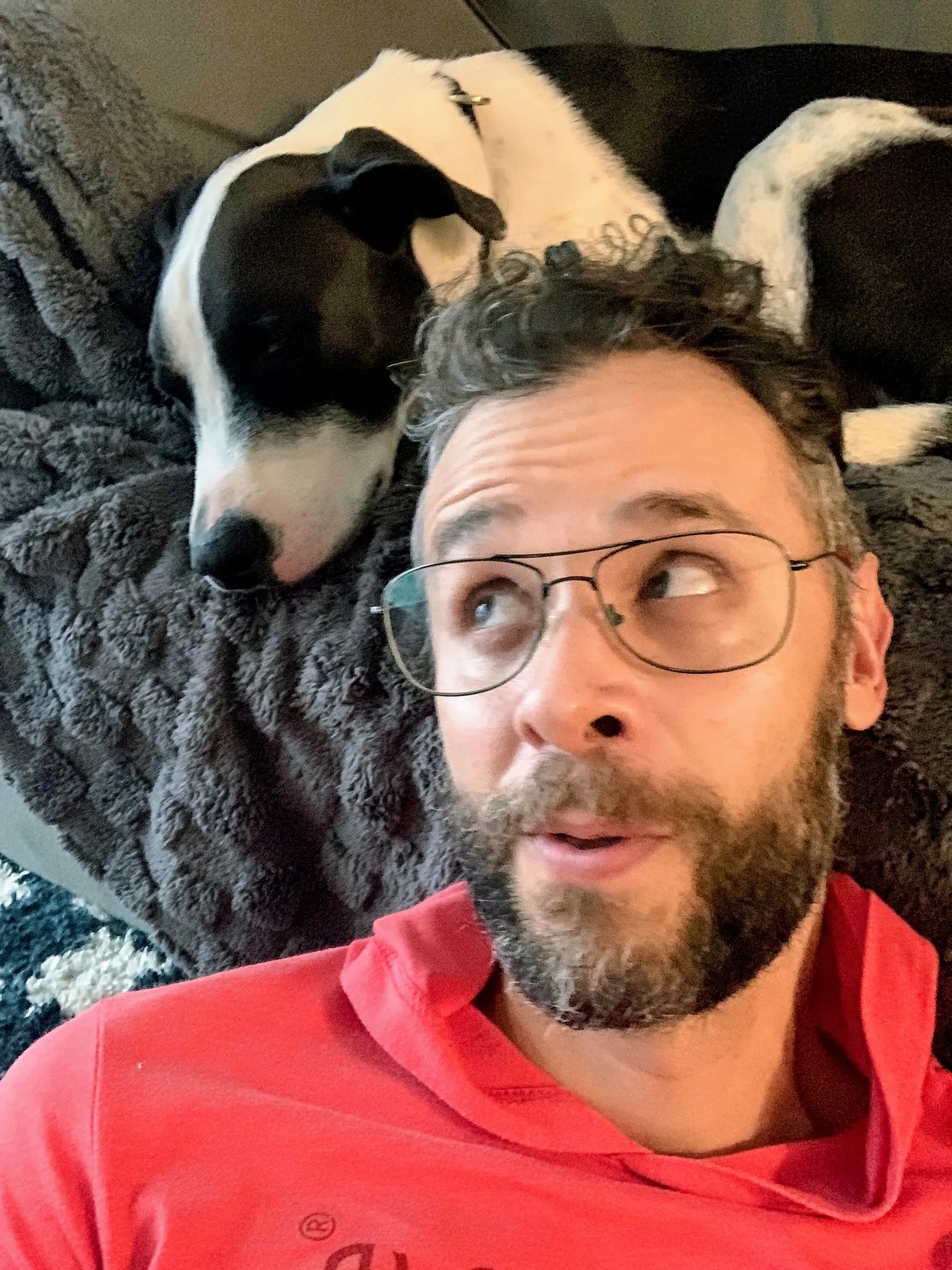 Selfie of Diego in a red hoodie glancing sideways at Forrest, a black and white dog curled up on a blanket just above his head