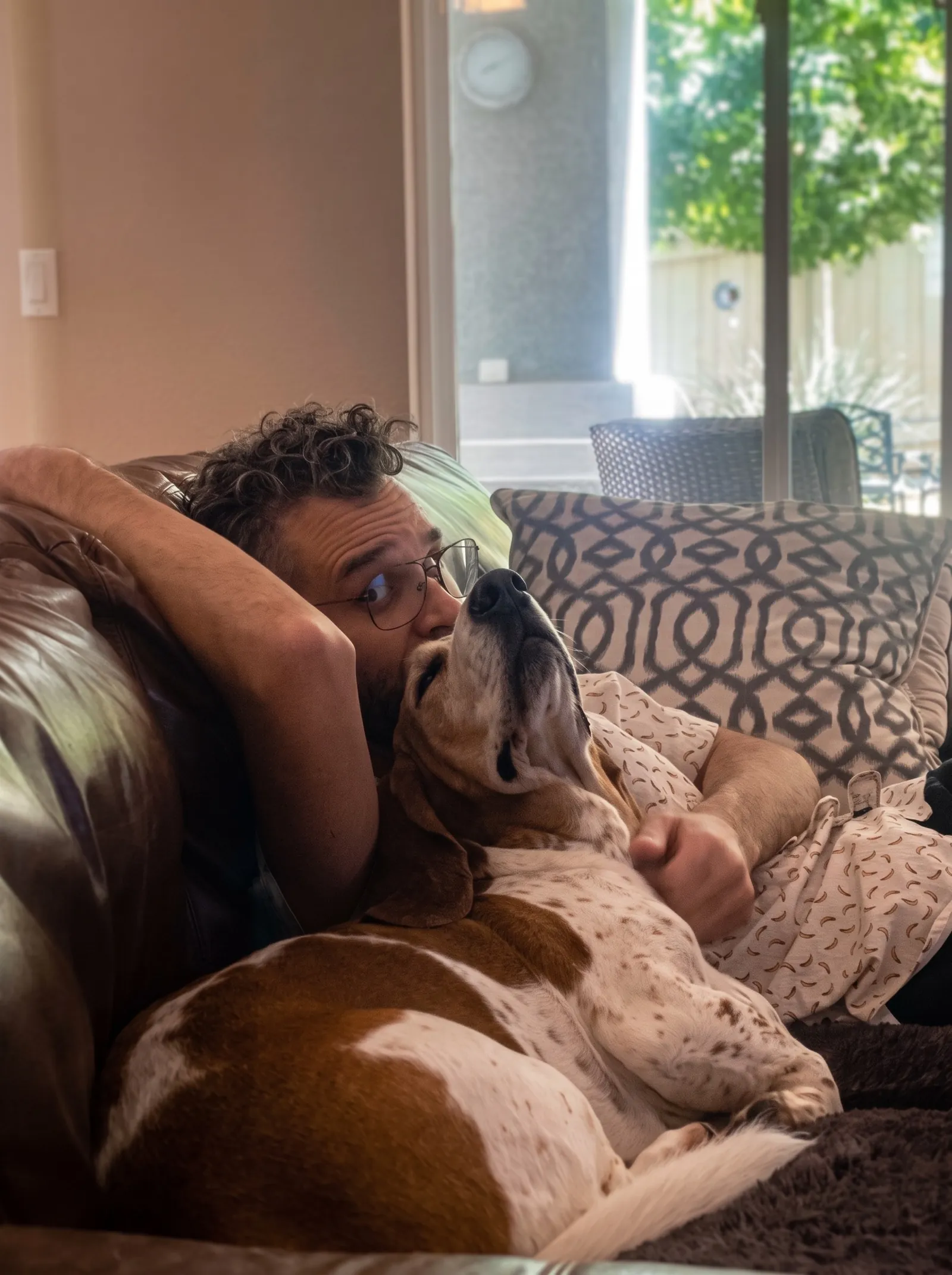 Diego lying on a leather couch with Xobee, a brown and white basset hound, stretched across his chest with her nose pointing up at his face